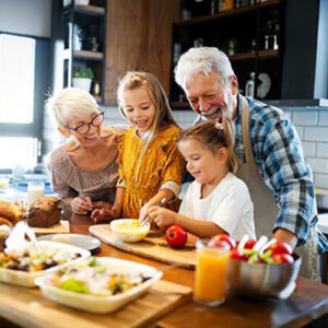 Older man and woman with two young girls preparing food together in a bright kitchen, smiling and enjoying the activity, with various dishes and drinks on the wooden counter in front of them.