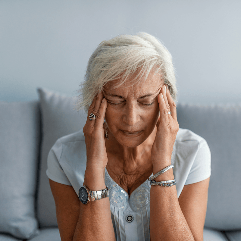 Older woman with short white hair sits on a couch, eyes closed, pressing her temples with her fingers, appearing to have a headache or feel stressed.