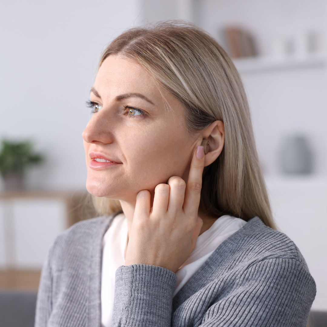 A woman with blonde hair touches her ear with her finger, looking thoughtful. She wears a gray sweater and sits indoors with blurred shelves and a plant in the background.