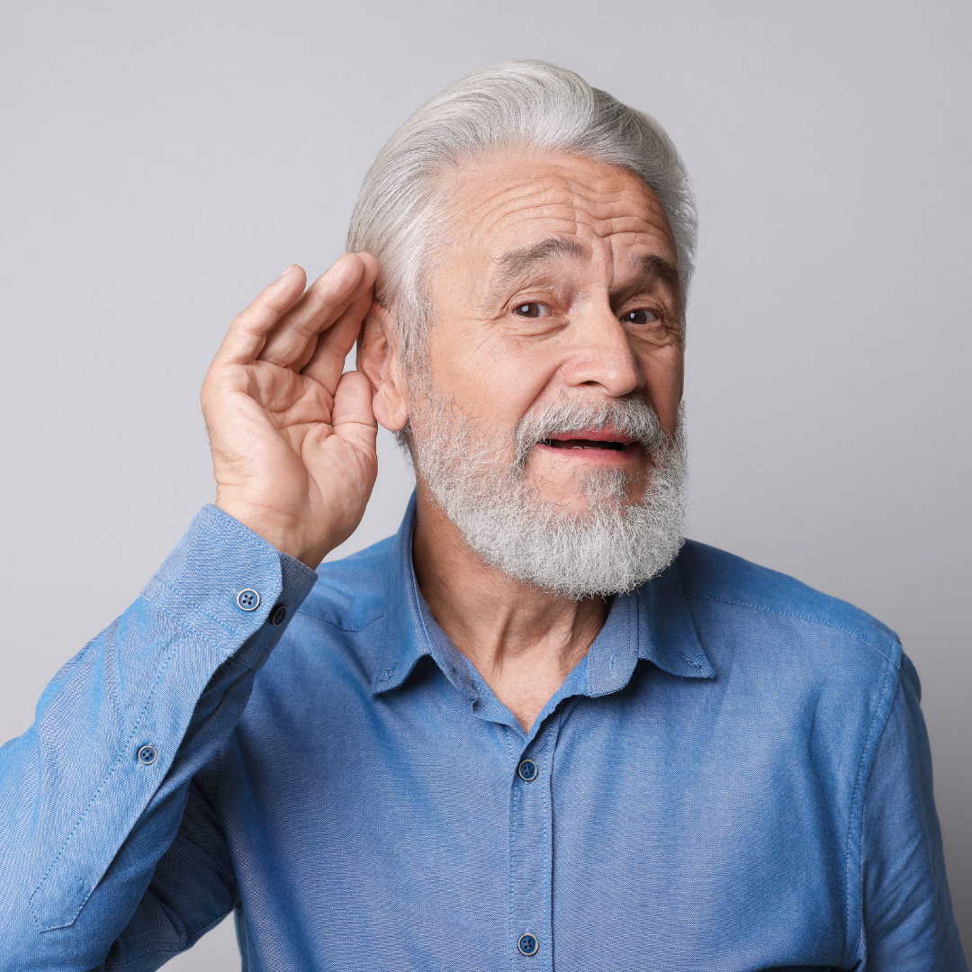 Older man with gray hair and beard wearing a blue shirt, holding his hand to his ear as if trying to hear better, against a plain gray background.