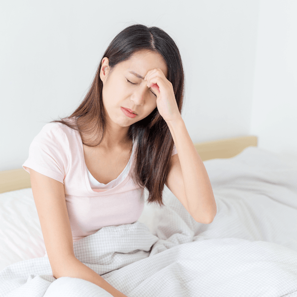 A woman sitting in bed with her eyes closed, holding her forehead in pain.